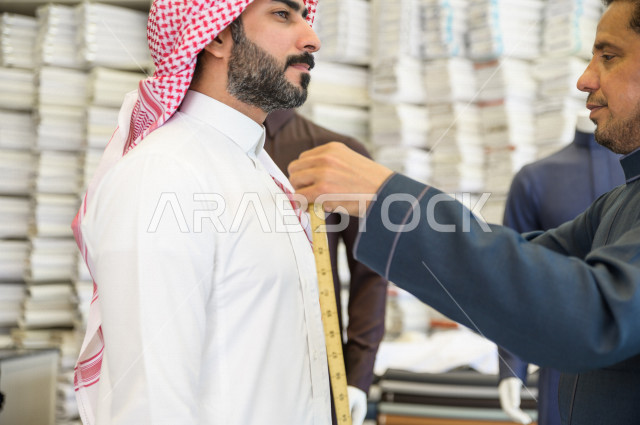 A young Saudi Gulf man working in the profession of sewing, he takes measurements of the customer's body in terms of length, width and waist using metric tape, a shop for sewing and tailoring men's clothes in Saudi Arabia, sewing skill and mastering the d