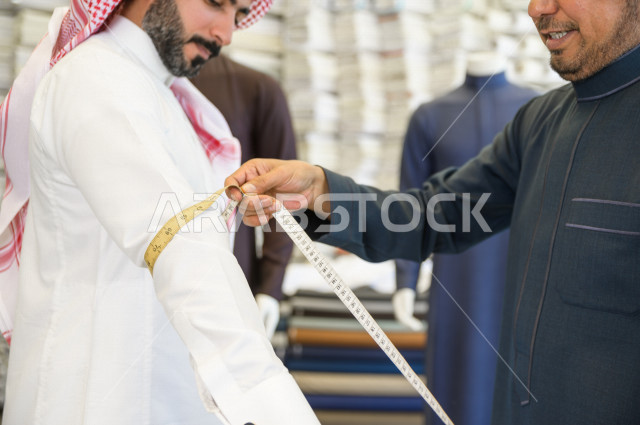 شاب سعودي خليجي يعمل في مهنة الخياطة ، يقوم بأخذ القياسات الخاصة بجسم الزبون من حيث الطول و العرض و الخصر باستخدام الشريط المتري ، محل خياطة و تفصيل الملابس الرجالي في السعودية ، مهارة الخياطة واتقان تفصيل الملابس الشبابية