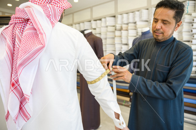 شاب سعودي خليجي يعمل في مهنة الخياطة ، يقوم بأخذ القياسات الخاصة بجسم الزبون من حيث الطول و العرض و الخصر باستخدام الشريط المتري ، محل خياطة و تفصيل الملابس الرجالي في السعودية ، مهارة الخياطة واتقان تفصيل الملابس الشبابية