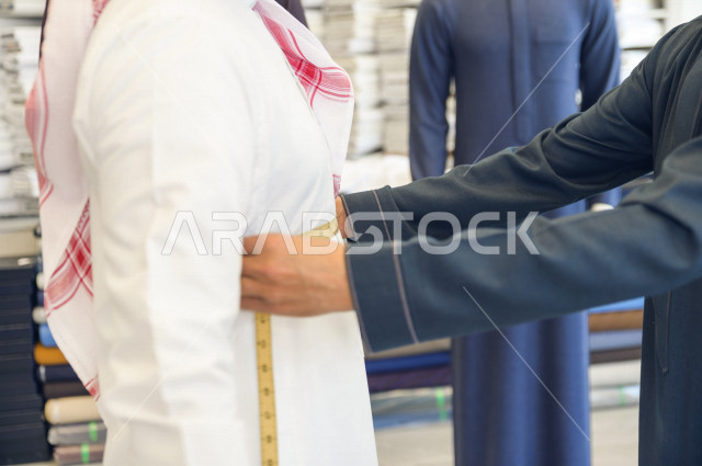 شاب سعودي خليجي يعمل في مهنة الخياطة ، يقوم بأخذ القياسات الخاصة بجسم الزبون من حيث الطول و العرض و الخصر باستخدام الشريط المتري ، محل خياطة و تفصيل الملابس الرجالي في السعودية ، مهارة الخياطة واتقان تفصيل الملابس الشبابية