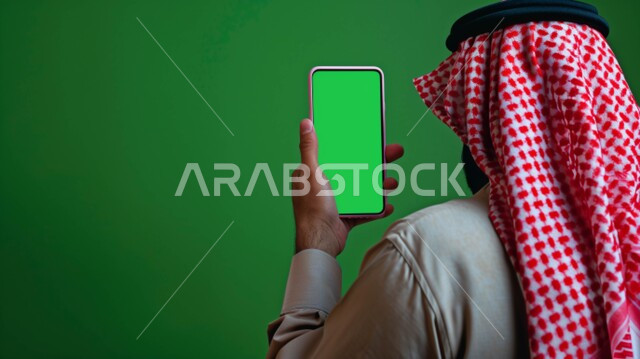 إدارة وانجاز المهام عبر الاونلاين, التواصل مع الأهل والأقارب عبر تطبيقات الجوال, استخدام الأجهزة والتقنيات الحديثة, بورتريه مقرب من الحلف لرجل عربي خليجي سعودي يرتدي الشماغ  والثوب التقليدي يحمل بيده هاتف محمول بشاشة خضراء فارغة, كروما, خلفية خضراء