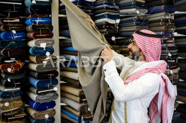 شاب سعودي خليجي يعمل في مهنة الخياطة ، اختيار اجود انواع الأقنشة لتفصيل الثوب السعودي ، محل خياطة و تفصيل الملابس الرجالي في السعودية ، مهارة الخياطة واتقان تفصيل الملابس الشبابية