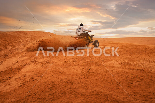 السماء المليئة بالغيوم فوق الرمال الذهبية في وقت غروب الشمس، مغامرات سفاري على الكثبان الرملية، أنشطة شبابية ممتعة في البيئة الصحراوية، صورة من الخلف لامرأة عربية خليجية سعودية ترتدي الزي الكاجوال وخوذة الحماية يقوم بقيادة دراجة الدفع الرباعي وسط الصحراء