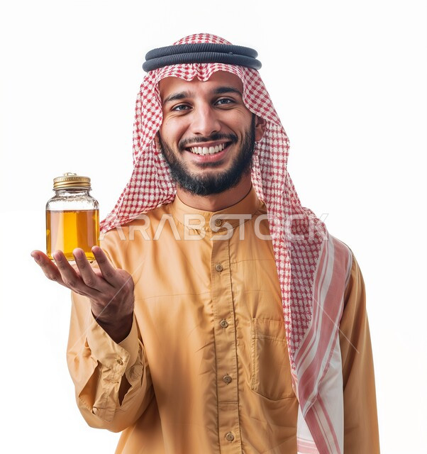 النظر إلى الكاميرا بايماءات السرور والسعادة، بورتريه مقرب لشاب عربي خليجي سعودي يرتدي الثوب التقليدي يحمل بيده عبوة زجاجية من عسل النحل الطازج، منتجات وطنية محلية سعودية، غذاء طبيعي صحي، خلفية بيضاء