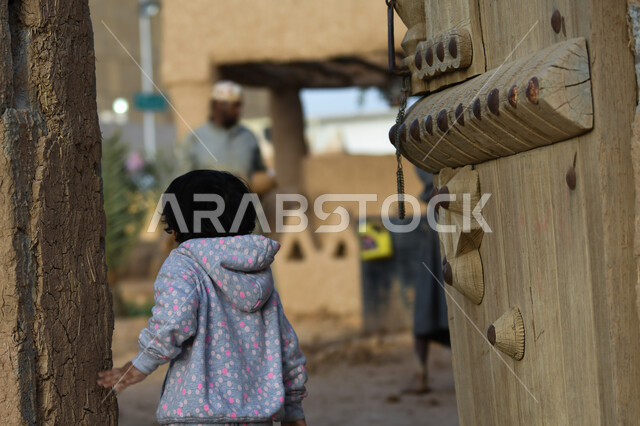 الأبواب الخشبية في المباني الطينية الشعبية، صورة من الخلف لطفلة عربية خليجية سعودية في احدى القرى القديمة، اسلوب الحياة التراثي الشعبي في المملكة العربية السعودية