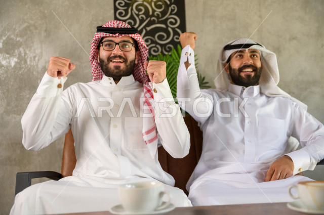 شابان عربيان سعوديان يجلسان داخل المقهى , مع تعابير وجه تدل على الفرحة والفوز , تحقيق اهداف