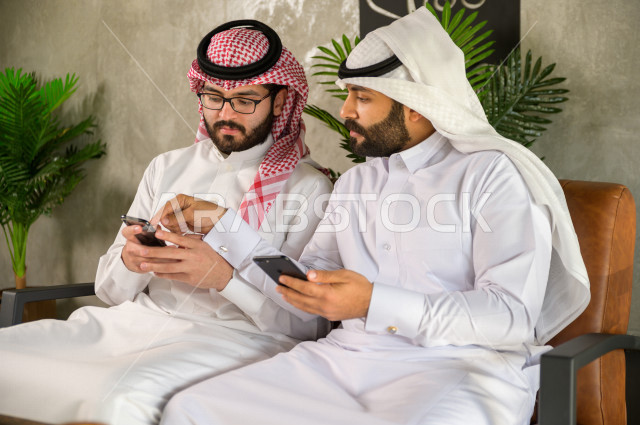 صديقان سعوديان يتبادلان المعلومات من الهاتف المحمول داخل المقهى , مناقشة عمل بواسطة الهاتف الذكي في كافيه
