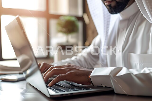 بيئة العمل المريحة في الشركات السعودية, مهن ووظائف مكتبية, صورة مقربة لرجل عربي خليجي سعودي يرتدي الغترة والثوب التقليدي ينظر إلى الكاميرا يقوم باستخدام الحاسب المحمول, إدارة وتنظيم الأعمال ضمن مقر الشركة، دمج التقنية والتكنولوجيا مع الحياة العملية
