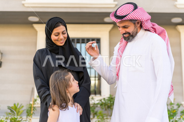 زوج سعودي خليجي يفاجئ عائلته بامتلاك منزل جديد ، يحمل الزوج بيده مفتاح المنزل الجديد ، شراء عقار جديد ، استقرار العائلة ،ايماءات وجه و يدين تدل على السعادة والانسجام ، منازل و عقارات