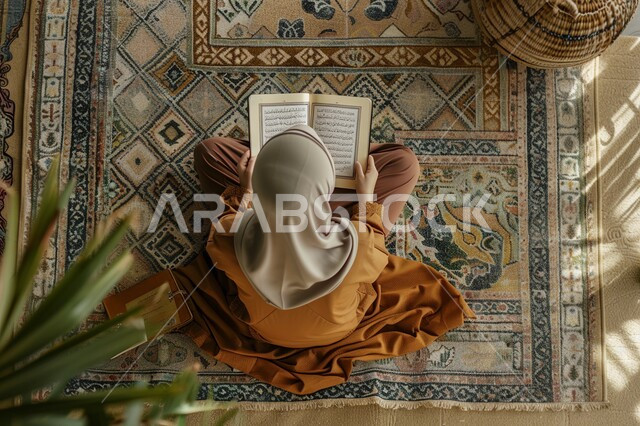الإكثار من ذكر الله تعالى والإجتهاد في العباده، تلاوة وقراءة القرآن الكريم، مفهوم الطمأنينه والخشوع، صورة من الاعلى لإمرأة عربية سعودية خليجية ترتدي الحجاب والعباءة تجلس على سجادة الصلاة تحمل بيدها المصحف الشريف، التقرب الى الله في شهر رمضان