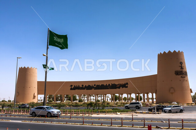 علم المملكة يرفرف فوق نصب الفاتحين التاريخي للمؤسس عبد العزيز في مدينة الرياض في النهار، الاهتمام بالتشجير والنباتات الخضراء في الاماكن الاثرية السياحية في المملكة العربية السعودية، معالم الرياض التراثية، فن الهندسة المعمارية والمباني الطينية القديمة