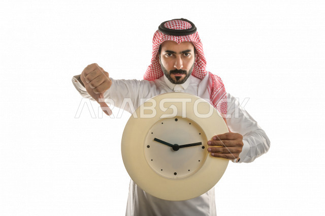 بورتريه لرجل سعودي خليجي غاضب و عابس بإيماءات وجه و يدين مختلفة  ، تعابير وجه و يدين مختلفة , يشير اصبع الإبهام الى الأسفل، يحمل بيده ساعة حائط  ، خلفية بيضاء