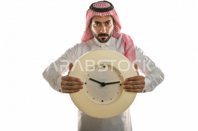 بورتريه لرجل سعودي خليجي  سعيد بإيماءات الوجه المختلفة  ، تعابير وجه و يدين مختلفة  ، يحمل بيده ساعة حائط  ، يشير اصبعه الى الساعة ،خلفية بيضاء