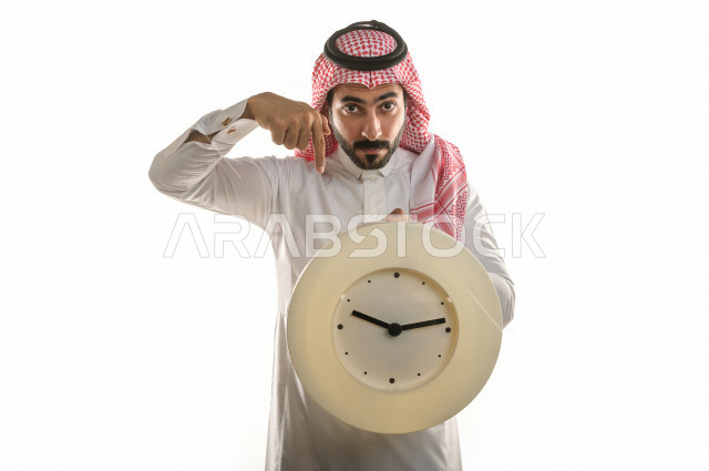بورتريه لرجل سعودي خليجي  سعيد بإيماءات الوجه المختلفة  ، تعابير وجه و يدين مختلفة  ، يحمل بيده ساعة حائط  ، يشير اصبعه الى الساعة ،خلفية بيضاء