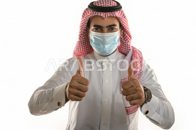 بورتريه لشاب سعودي يقوم بوضع الكمامة ، اتباع اجراءات السلامة بوضع الكمامة بشكل صحيح ، تجنب انتشار العدوى ، رفع اصبع الإبهام   ، خلفية بيضاء