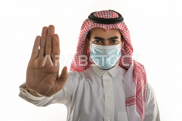 بورتريه لشاب سعودي يرتدي الكمامة ، يرفع يده ليتجنب المصافحة واقتراب الاخرين منه ، تجنب انتشار العدوى ، الحفاظ على المسافات ، خلفية بيضاء