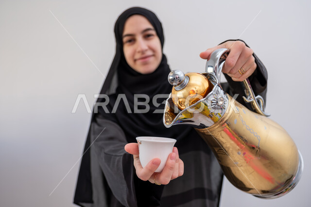 كرم الضيافة وحسن الاستقبال في السعودية, بورتريه مقرب لشابة عربية خليجية سعودية مبتسمة ترتدي العباءة والحجاب تقوم بصب القهوة الخليجية, تقديم القهوة  العربية الأصيلة في الأعياد, أصالة عادات وتقاليد المجتمع السعودي في المناسبات الاسلامية, خلفية بيضاء