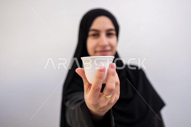 تقديم القهوة العربية في المناسبات الاسلامية, بورتريه مقرب لشابة عربية خليجية سعودية مبتسمة ترتدي العباءة والحجاب تمسك بيدها فنجان القهوة, الاستمتاع بالمشروبات الساخنة, أصالة العادات والتقاليد في المملكة العربية السعودية, خلفية بيضاء