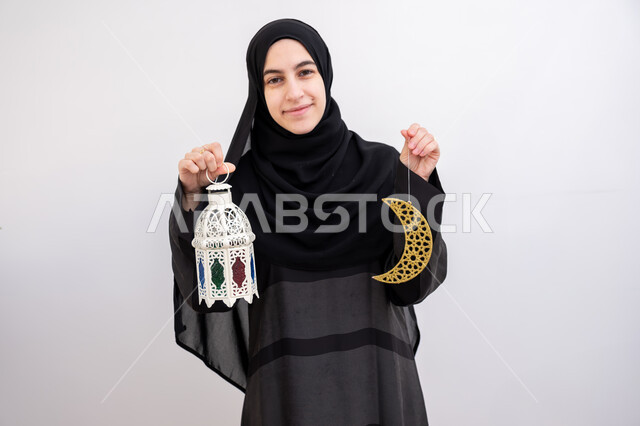  زينة المناسبات والأعياد الاسلامية, بورتريه لشابة عربية خليجية سعودية مبتسمة ترتدي الحجاب والعباءة تمسك بيدها هلال وفانوس مزخرف بإيماءات السرور والسعادة, بهجة وفرحة المسلمين بقدوم شهر الخير,الاستعداد لاستقبال شهر رمضان المبارك, خلفية بيضاء