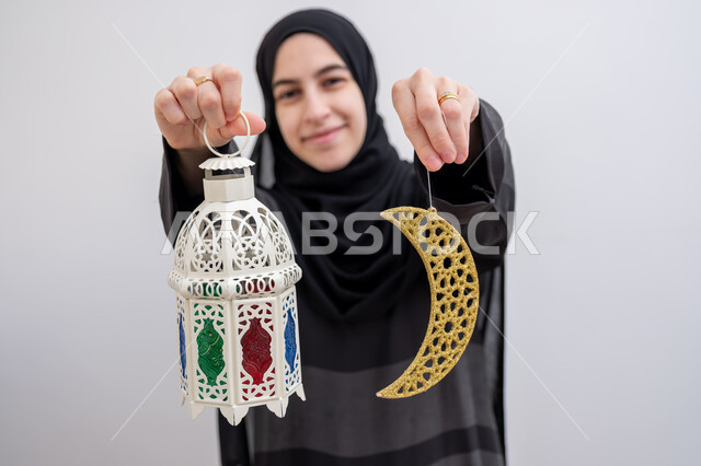 الاستعداد لاستقبال شهر رمضان المبارك, زينة المناسبات والأعياد الاسلامية, بورتريه مقرب لشابة عربية خليجية سعودية مبتسمة ترتدي الحجاب والعباءة تمسك بيدها هلال وفانوس مزخرف بإيماءات السرور والسعادة, بهجة وفرحة المسلمين بقدوم شهر الخير, خلفية بيضاء