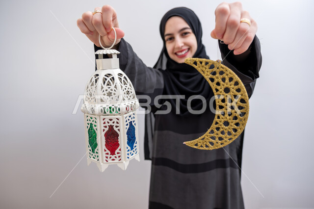  زينة المناسبات والأعياد الاسلامية, بورتريه لشابة عربية خليجية سعودية مبتسمة ترتدي الحجاب والعباءة تمسك بيدها هلال وفانوس مزخرف بإيماءات السرور والسعادة, بهجة وفرحة المسلمين بقدوم شهر الخير,الاستعداد لاستقبال شهر رمضان المبارك, خلفية بيضاء