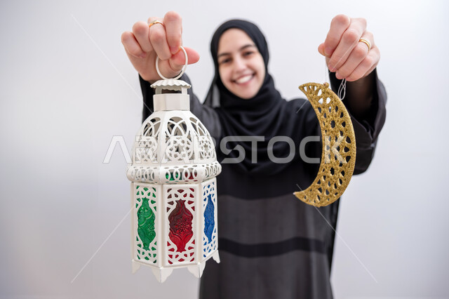 The joy and happiness of Muslims with the arrival of the month of goodness, preparation to welcome the holy month of Ramadan, decorations for Islamic occasions and holidays, portrait of a smiling Saudi Gulf Arab young woman wearing the hijab and abaya, holding in her hand a crescent moon and a lantern decorated with gestures of pleasure and happiness, white background