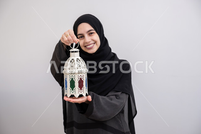 زينة وفوانيس الأعياد والمناسبات الاسلامية في المملكة العربية السعودية, بورتريه مقرب لشابة عربية خليجية سعودية محجبة مبتسمة ترتدي العباءة الرمادية تمسك بيدها فانوس ملون, الاستعداد لاستقبال شهر رمضان المبارك بإيماءات السرور, خلفية بيضاء