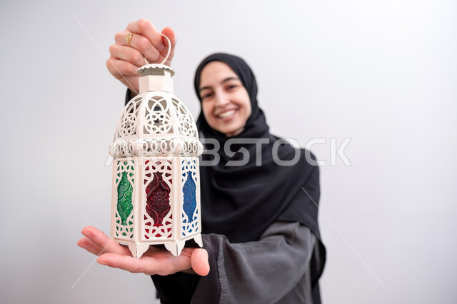فوانيس وزينة الأعياد والمناسبات الاسلامية في المملكة العربية السعودية, بورتريه مقرب لشابة عربية خليجية سعودية محجبة مبتسمة ترتدي العباءة الرمادية تمسك بيدها فانوس ملون, الاستعداد لاستقبال شهر رمضان المبارك بإيماءات السرور, خلفية بيضاء