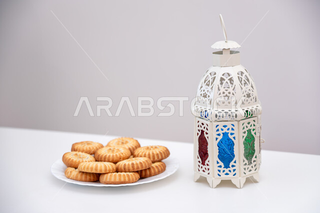 حلويات شعبية تقليدية لذيذة، زينة وفوانيس المناسبات الإسلامية والدينية، كرم الضيافة وحسن الاستقبال في العيد، أجواء الاحتفال بالأعياد في السعودية، طبق حلى المعمول المحشو بالتمر بجانب فانوس أبيض معدني مزخرف