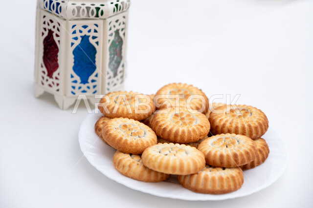 طبق حلى المعمول المحشو بالتمر بجانب فانوس أبيض معدني مزخرف، حلويات شعبية تقليدية لذيذة، زينة وفوانيس المناسبات الإسلامية والدينية، كرم الضيافة وحسن الاستقبال في العيد، أجواء الاحتفال بالأعياد في السعودية