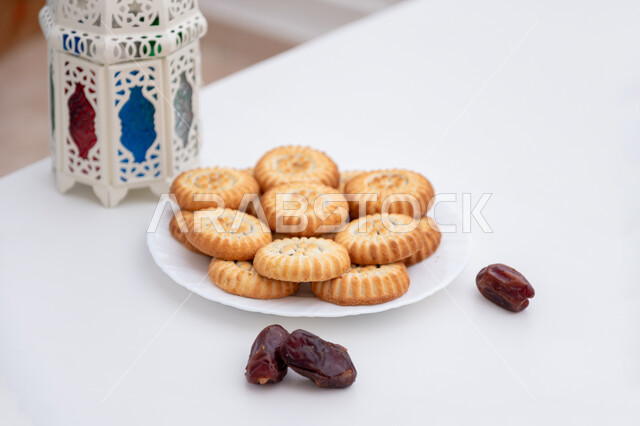 كرم الضيافة وحسن الاستقبال في العيد، أجواء الاحتفال بالأعياد في السعودية، طبق حلى المعمول المحشو بالتمر بجانب فانوس أبيض معدني مزخرف، حلويات شعبية تقليدية لذيذة، زينة وفوانيس المناسبات الإسلامية والدينية