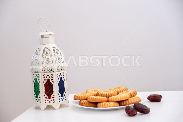  زينة وفوانيس المناسبات الإسلامية والدينية، كرم الضيافة وحسن الاستقبال في العيد، أجواء الاحتفال بالأعياد في السعودية، طبق حلى المعمول المحشو بالتمر بجانب فانوس أبيض معدني مزخرف، حلويات شعبية تقليدية لذيذة