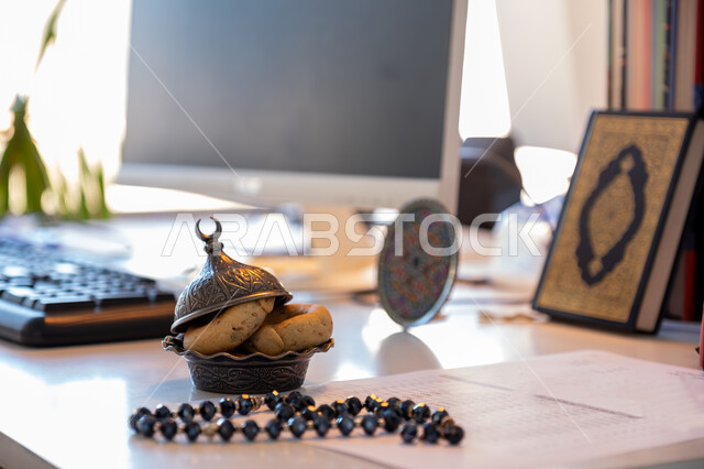  سبحة مصنوعه من الخرز لذكر الله تعالى, الالتزام والتمسك بالذكر، كعك العيد الشهي, ضيافة المناسبات والأعياد في المملكة العربية السعودية, صورة مقربة لطبق من الحلويات الشعبية على المكتب بجانب المصحف الشريف, الأقراص المخبوزة الطازجة