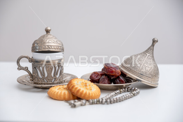 Delicious Eid cakes with a bowl full of dates, hosting events and holidays in the Kingdom of Saudi Arabia, a close-up of a plate of popular sweets next to a cup of coffee, a rosary made of beads for the remembrance of God Almighty, commitment and adherence to the remembrance, white background