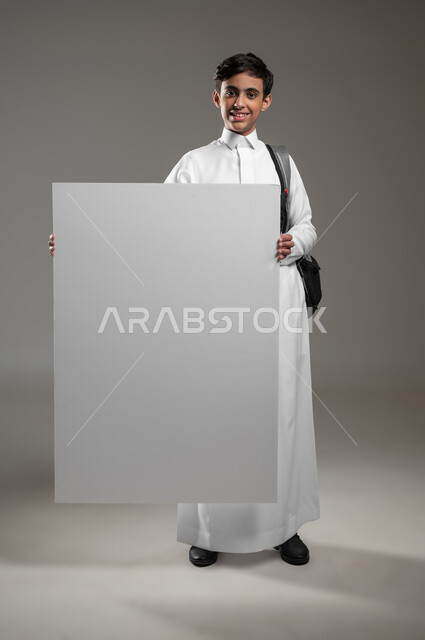A large white rectangular mockup, announcing the beginning of a new academic year in advanced Saudi schools, a portrait of a Saudi Gulf Arab student wearing a traditional dress and a backpack holding an empty billboard in his hand, expressions of pleasure and happiness, full body image, gray background, sensitive use, sensitive content