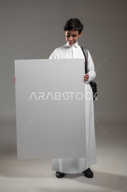 الاعلان عن بداية عام دراسي جديد في المدارس السعودية المتقدمة، موك اب مستطيل ابيض كبير، بورتريه لطالب عربي خليجي سعودي مبتسم يرتدي الثوب التقليدي وحقيبة الظهر يحمل بيده لوح فارغ، تعابير السرور والسعادة، صورة الجسم بالكامل، خلفية رمادية، محتوى حساس، استخدام حساس
