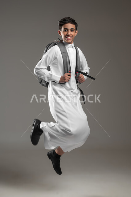 الاستعداد لبدء عام دراسي جديد، المدارس السعودية المتطورة، بورتريه لطالب عربي خليجي سعودي مبتسم يرتدي الثوب التقليدي وحقيبة الظهر المدرسية يقفز عاليا بتعابير وايماءات الحماس والسرور للذهاب الى المدرسة، صورة الجسم بالكامل،  خلفية رمادية، محتوى حساس، استخدام حساس