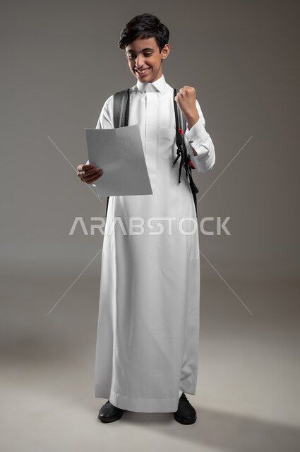 ايماءات وتعابير اليد تدل على الفوز والنجاح، بورتريه لطالب عربي خليجي سعودي مبتسم يرتدي الثوب التقليدي وحقيبة الظهر المدرسية يحمل بيده مجموعة أوراق، اجتياز الاختبارات بتفوق ودرجات عليا، صورة الجسم بالكامل، خلفية رمادية، محتوى حساس، استخدام حساس