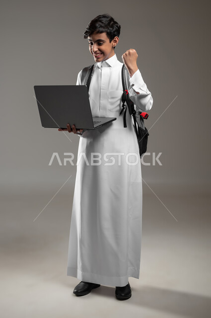 دمج العلم مع التقنية والتكنولوجيا، ايماءات الفرح لصدور نتائج الاختبارات النهائية اونلاين، بورتريه لطالب عربي خليجي سعودي مبتسم يرتدي الثوب التقليدي يحمل بيده الحاسوب المحمول بتعابير النجاح والحماس، صورة لكامل الجسم، خلفية رمادية، استخدام حساس، محتوى حساس