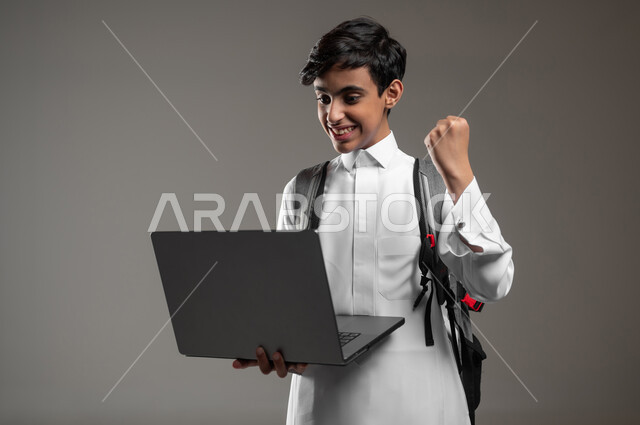 ايماءات الفرح لصدور نتائج الاختبارات النهائية اونلاين، بورتريه مقرب لطالب عربي خليجي سعودي مبتسم يرتدي الثوب التقليدي يحمل بيده الحاسوب المحمول بتعابير النجاح والحماس، دمج العلم مع التقنية والتكنولوجيا، خلفية رمادية، استخدام حساس، محتوى حساس