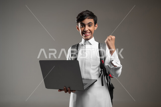النظر الى الكاميرا بايماءات الفرح لصدور نتائج الاختبارات النهائية اونلاين، بورتريه مقرب لطالب عربي خليجي سعودي مبتسم يرتدي الثوب التقليدي يحمل بيده الحاسوب المحمول بتعابير النجاح والحماس، دمج العلم مع التقنية والتكنولوجيا، خلفية رمادية، محتوى حساس، استخدام حساس