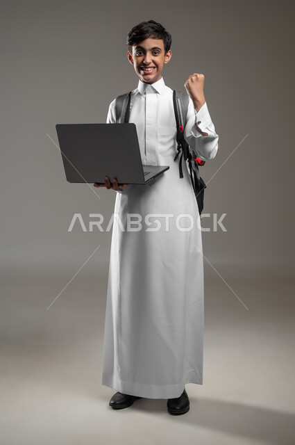 Issuing final exam results online, integrating science with technology and technology, portrait of a smiling Saudi Arabian Gulf student wearing a traditional dress holding a laptop in his hand with expressions of success and enthusiasm, full body photo, gray background, sensitive content, sensitive use