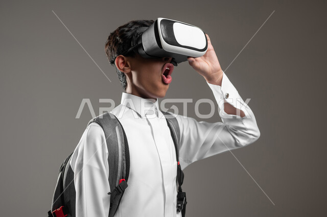 محاكاة العالم الخيالي بتعابير الصدمة والذهول، الدراسة عبر منصة تعليمية للتعلم عن بعد، بورتريه مقرب لطالب عربي خليجي سعودي يرتدي الثوب التقليدي وحقيبة الظهر يضع نظارة الفي آر، نشاطات الدراسة باستخدام الواقع الافتراضي، خلفية رمادية، محتوى حساس، استخدام حساس