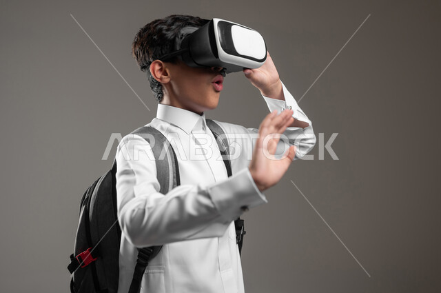 محاكاة العالم الافتراضي بتعابير الدهشة، الدراسة عبر منصة تعليمية للتعلم عن بعد، بورتريه مقرب لطالب عربي خليجي سعودي يرتدي الثوب التقليدي وحقيبة الظهر المدرسية يضع نظارة الفي آر يقوم بصد شيء ما، خلفية رمادية، محتوى حساس، استخدام حساس