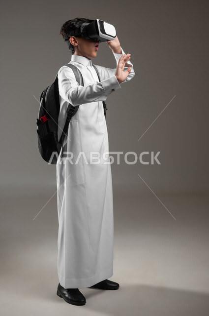 محاكاة الواقع الافتراضي بتعابير الدهشة، الدراسة عبر منصة تعليمية للتعلم عن بعد، بورتريه لطالب عربي خليجي سعودي يرتدي الثوب التقليدي وحقيبة الظهر المدرسية يضع نظارة الفي آر يقوم بصد شيء ما، صورة الجسم بالكامل، خلفية رمادية، محتوى حساس، استخدام حساس