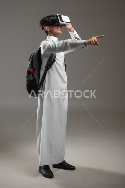 Pointing to something and simulating the imaginary world with expressions of pleasure, a portrait of a Saudi Arabian Gulf student wearing a traditional dress and a school backpack, wearing VR glasses, a study activity using virtual reality, full body image, gray background, sensitive content, sensitive use