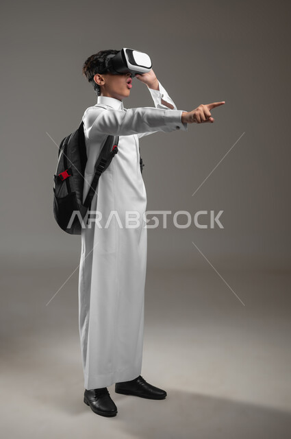 الاشارة الى شيء ما ومحاكاة العالم الخيالي بتعابير الدهشة، بورتريه لطالب عربي خليجي سعودي يرتدي الثوب التقليدي وحقيبة الظهر المدرسية يضع نظارة الفي آر، نشاط دراسي باستعمال الواقع الافتراضي، صورة الجسم بالكامل، خلفية رمادية، محتوى حساس، استخدام حساس