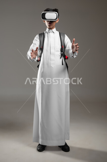 محاولة الإمساك بشيء ما، نشاط دراسي باستعمال الواقع الافتراضي، الدراسة عبر منصة تعليمية للتعلم عن بعد، بورتريه لطالب عربي خليجي سعودي يرتدي الثوب التقليدي وحقيبة الظهر المدرسية يضع نظارة الفي آر، صورة الجسم بالطول الكامل، خلفية رمادية، محتوى حساس، استخدام حساس