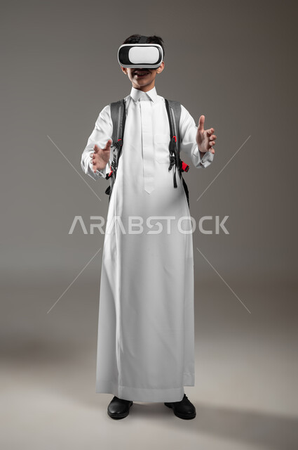 محاولة الإمساك بشيء ما في العال الخيالي، نشاط دراسي باستعمال الواقع الافتراضي، الدراسة عبر منصة تعليمية للتعلم عن بعد، بورتريه لطالب عربي خليجي سعودي يرتدي الثوب التقليدي وحقيبة الظهر يضع نظارة الفي آر، صورة لكامل الجسم، خلفية رمادية، محتوى حساس، استخدام حساس