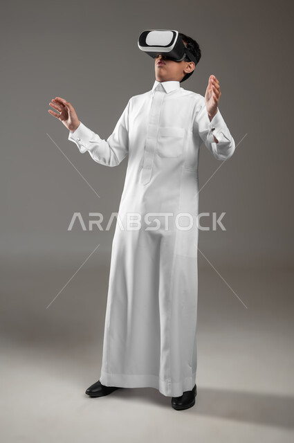 Expressions and gestures indicating integration and astonishment, a portrait of a surprised Saudi Gulf Arab boy wearing a traditional dress, watching movies through 3D virtual reality glasses, simulating the imaginary world and interacting with it, full picture, gray background, sensitive content, sensitive use